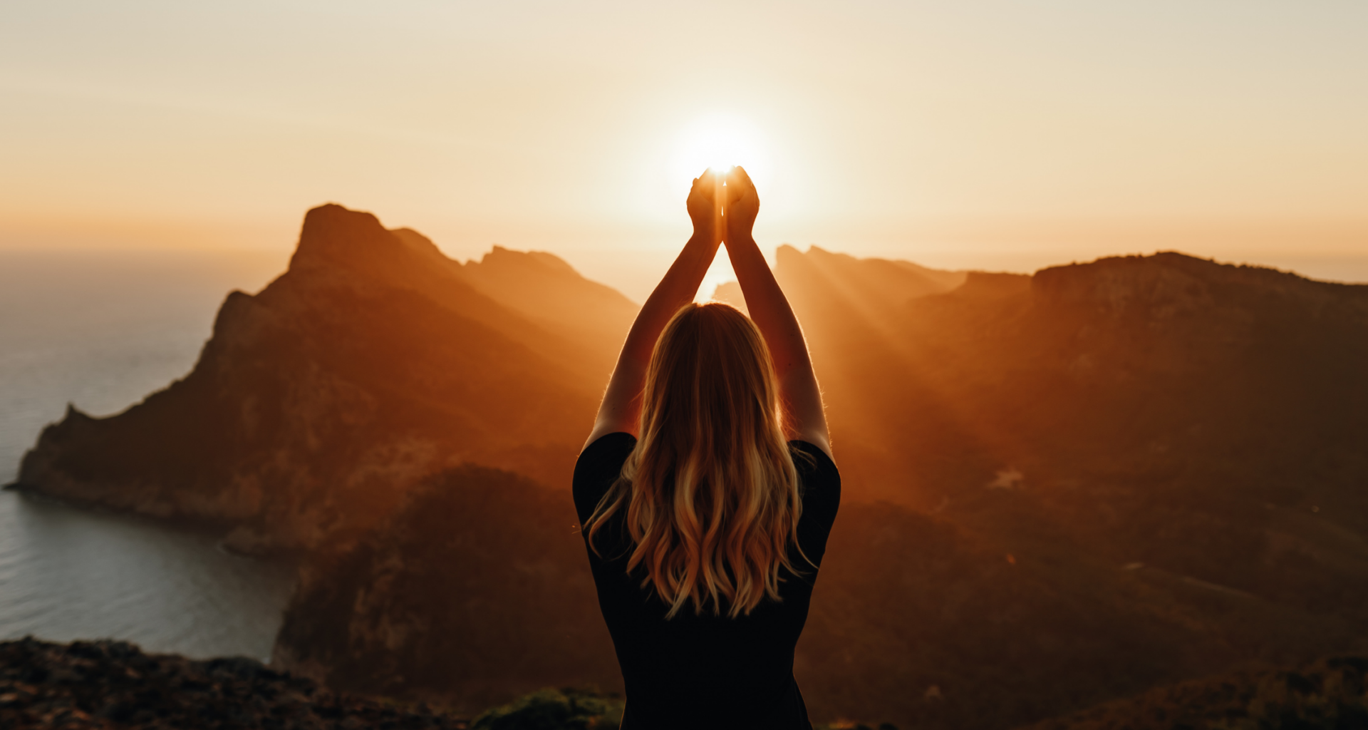young woman with arms in the air posing to look like she's holding the sunlight that is peeking over the mountains