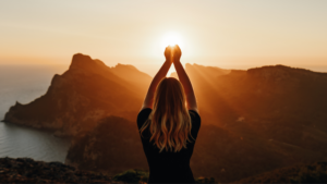 young woman with arms in the air posing to look like she's holding the sunlight that is peeking over the mountains